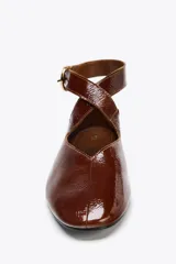 Zapato tipo bailarina color marrón, con efecto acharolado, punta redonda y cierre mediante tira con hebilla al tobillo.