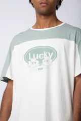 Remera color beige con mangas y parte superior en verde militar. Estampado frontal con texto "Lucky" y diseño de flores con rostro.