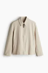 Campera color beige de tejido suave con cuello, cierre frontal, puños ajustables con botón a presión y bolsillos insertados al bies. Corte estándar y mangas forradas.