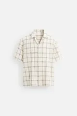 Camisa de manga corta color crudo con estampado de cuadros en tonos grises y marrones. Tiene cuello bowling, corte relaxed fit, aberturas laterales en el bajo y cierre frontal con botones.