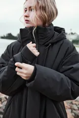 Anorak largo negro resistente al agua y al viento, con aislamiento térmico, cuello alto con capucha ajustable, bolsillos con cremallera, acolchado interior, tirantes elásticos y cremalleras laterales en el bajo.