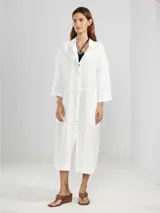 Camisa larga blanca de lino italiano, con cuello clásico, mangas 3/4, silueta fluida, dobladillo curvado en las costuras laterales y bolsillos discretos.