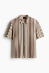 Camisa de manga corta con cuello inglés, botones adelante y bajo recto. Tejido de punto pointelle con estilo crochet y diseño a rayas verticales en tonos beige, marrón y blanco.