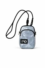 Cartera pequeña de color plateado brillante, con un bolsillo frontal con cierre y un parche negro con el logo "IND" en blanco. Tiene una correa fina negra ajustable.