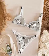 Conjunto de bikini blanco con estampado floral en tonos rosa y negro. El corpiño es de corte triangular con nudo central y breteles finos. La bombacha es vedetina con detalle de nudos en los laterales.