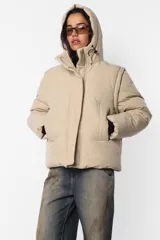Campera puffer beige de corte oversize, confeccionada en textil impermeable. Cuenta con capucha forrada con tancas y elástico, cuello alto, cierre frontal con cartera y abrojo, bolsillos ribete con cierre, mangas desmontables con mitones y cintura ajustable con elástico y tancas. Incluye cinta gross estampada para transportarla al hombro y bolsillo interno con cierre invisible.