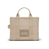 Tote bag de lona color beige con la inscripción "THE TOTE BAG MARC JACOBS" en el frente. Incluye cierre de cremallera, asas superiores y una correa de hombro ajustable y extraíble de color negro.