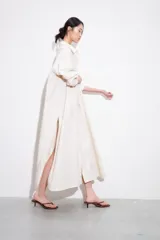 Vestido camisero largo de manga larga, color blanco con finas rayas verticales en tono claro, con cuello solapa y abotonadura frontal. Posee un calce amplio y bolsillos laterales.