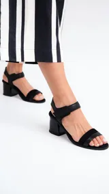 Sandalias de cuero negro con dos tiras, una sobre los dedos y otra al tobillo con cierre de velcro. Tacón bloque bajo.