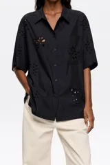 Camisa negra de manga corta con cuello clásico y cierre frontal con botones. Presenta bordado inglés con motivos florales. Tiene un bolsillo de parche en el pecho.