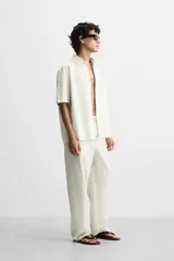 Pantalón relaxed fit de color beige con finas rayas verticales, confeccionado en una mezcla de algodón y cáñamo. Presenta cintura elástica, bolsillos laterales, un bolsillo trasero de plastrón y cierre frontal con botones.