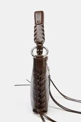 Bolso de hombro color gris topo, con formato rectangular y asa corta. Presenta dos bolsillos frontales con cierre de cremallera y detalle de tiras colgantes. El asa es de cuero trenzado con herrajes metálicos.