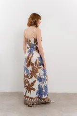 Vestido largo con estampado de hojas en tonos verdes y marrones sobre fondo blanco. Tiene breteles finos, escote en V y detalle de lazo en el frente. La falda es amplia y con vuelo.