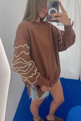 Buzo de algodón color blanco roto, de corte holgado, con puños y tajos laterales. Presenta un bordado artesanal en relieve de líneas orgánicas en color marrón caramelo sobre las mangas.