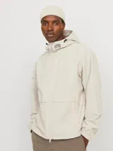 Chaqueta shell color beige claro, con capucha ajustable y cierre frontal con cremallera. Presenta logo de la marca en el pecho, mangas largas con puños elásticos, bolsillos laterales y corte recto.
