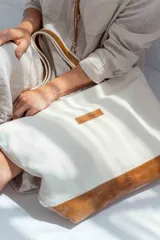 Bolso tipo tote de lona color crudo con base y asas de cuero color marrón.