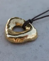 Collar con dije dorado irregular con agujero central, colgando de cordón negro ajustable.