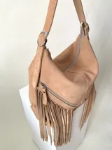 Bolso tipo hobo color tostado, hecho de cuero con flecos largos en el frente y dos bolsillos con cierre. Tiene una manija regulable con hebillas metálicas.