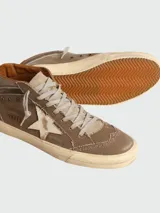 Zapatillas de media caña marca Golden Goose, modelo Mid-Star. Confeccionadas en lona color marrón con efecto desgastado, presentan una estrella de piel blanca en el lateral y detalles de brogue en la puntera y el talón. Cuentan con cordones grises y suela de goma con acabado envejecido.