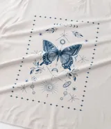 Remera color beige de corte alargado, cuello redondo y mangas cortas. Estampado frontal con una mariposa azul rodeada de elementos celestiales.