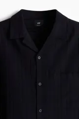 Camisa negra de manga corta con cuello resort, tapeta lisa, bolsillo superior y canesú en la espalda.