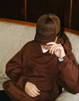 Gorra trucker marrón con frente de tela y parte trasera de rejilla. Presenta un bordado 3D con un diseño abstracto en el frente.