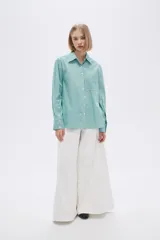 Camisa a rayas verticales verdes y blancas, de corte holgado, manga larga con puños abotonados, cuello clásico y bolsillo de parche en el pecho.