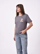 Remera blanca de algodón con cuello redondo y manga corta. Presenta un pequeño bordado en el pecho izquierdo con una figura que parece ser una persona acostada en una tabla de surf o flotador.