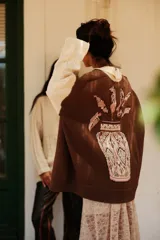 Buzo de punto en color marrón con cuello polo blanco y mangas largas blancas con textura de ochos. Presenta un diseño gráfico de gran tamaño en la espalda en tonos rosado, blanco y negro.