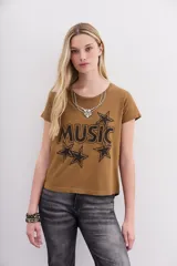 Remera color camel de mangas cortas confeccionada en jersey de algodón, de calce al cuerpo y escote redondo en punto rib a tono. Cuenta con una estampa central al agua de efecto desgastado con la palabra "Music" y estrellas, y un aplique de cadenas y piedras que simula ser un collar.