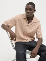 Camisa de manga corta Jack & Jones con cuello resort y cierre de botones. Diseño con estampado de espiga en tonos marrones y blancos. Confeccionada en tejido dobby 100% algodón.