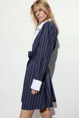 Vestido corto estilo camisero a rayas verticales azul marino y blancas, con cuello y puños blancos. Tiene cierre frontal con botones y lazo para atar en la cintura.
