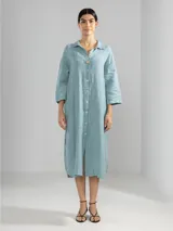Camisa larga celeste confeccionada en lino, con cuello clásico, mangas 3/4, silueta fluida, dobladillo curvado en las costuras laterales y bolsillos discretos.