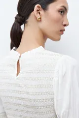 Blusa de color crudo con cuello alto fruncido y pechera con nido de abeja. Cuenta con mangas largas con detalles elásticos y puños con volados, y un bajo de corte peplum.