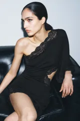 Vestido negro de escote asimétrico con un solo hombro y manga larga ancha. Presenta detalles de encaje negro en el escote, el ruedo asimétrico y un corte 'cut out' en el lateral del torso.