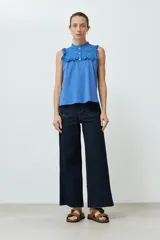Top blanco sin mangas, con cuello alto y volados en los hombros. Presenta detalles de broderie en el cuello y el pecho, y cierre de botones en la parte superior delantera.