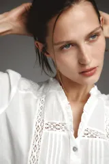 Camisa blanca de cuello subido y manga larga, con detalle de entredós combinados a tono y cierre frontal con botones.