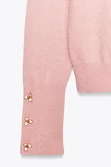 Sweater rosa de tejido de punto con cuello redondo y mangas largas. Detalle de botones dorados en relieve con forma de flor en los puños.