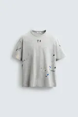 Camiseta gris con cuello redondo y mangas cortas. Tiene un corte relaxed fit y un estampado de salpicaduras de pintura en varios colores.