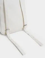 Mochila pequeña de cuero sintético color blanco con bolsillo frontal con solapa y cierre superior. Incluye un charm colgante de moño a rayas rojas y blancas con dije de corazón plateado.