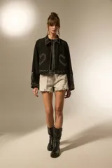 Campera cropped de jean color negro, con cuello camisero, cierre frontal y detalles de tachas plateadas en el cuello, hombros, mangas y ruedo. Presenta un diseño de tachas en forma de corazón en el pecho.