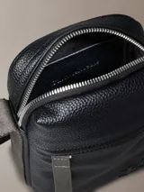 Cartera estilo reportera de Calvin Klein confeccionada en cuero sintético granulado negro. Posee un compartimento principal y un bolsillo frontal con cierre de cremallera, además del monograma de la marca grabado en relieve. Incluye una correa ajustable de color verde oliva.