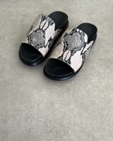 Sandalias chatas color beige con base negra y detalle de bordado blanco en la tira superior.