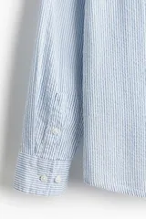 Camisa de hombre de corte holgado, confeccionada en tela de sirsaca a rayas verticales celestes y blancas. Tiene cuello inglés, tapeta clásica con botones, bolsillo de parche en el pecho, mangas largas con puños abotonados y bajo redondeado.