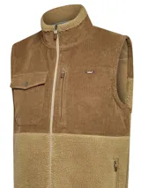 Chaleco beige con diseño de mitad sherpa y mitad cotelé, cuello alto y cierre frontal con cremallera. Incluye tres bolsillos con cremallera y un bolsillo con botón.