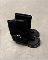 Botas negras de caña media, estilo sandalia-bota, con diseño tipo chinela en la parte delantera (dedo separado) y caña de tela suave y holgada. Presentan una tira ajustable con hebilla metálica plateada alrededor del tobillo.