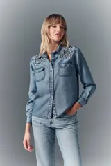 Camisa de denim celeste con apliques de piedras plateadas en los hombros, corte recto, cuello camisero, botones metálicos y bordado Rapsodia en la espalda.