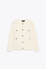 Chaqueta color crema de tejido de punto liso, con cuello redondo, manga larga, bolsillos delanteros de plastrón y cierre frontal con botones dorados.