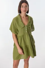 Vestido corto color verde oliva de lino, con cuello polo, mangas cortas y falda escalonada. Presenta detalle de puntilla en cuello y mangas, y cierre frontal con botones.