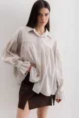 Camisa gris a rayas verticales blancas, de corte amplio, cuello alto con lazo y mangas largas abullonadas con puños elásticos.